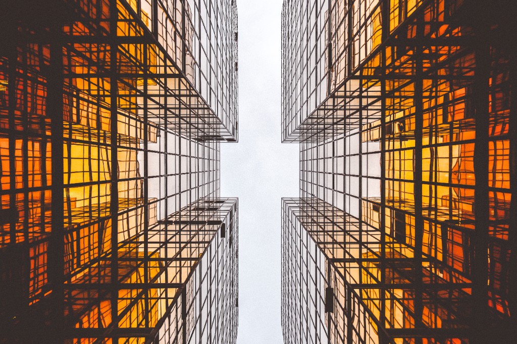 Looking up at four glass skyscrapers converging toward a bright sky, with warm amber light in the windows.